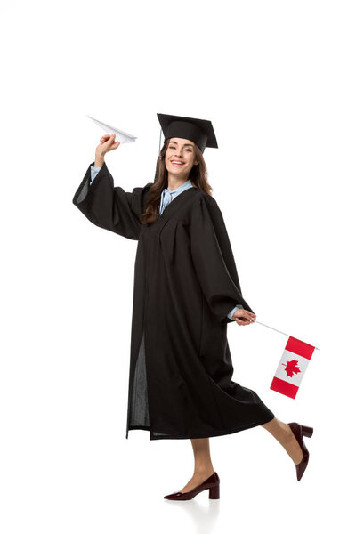 happy female student in academic gown holding canadian flag and paper plane isolated on white