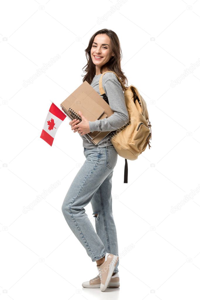 happy female student with backpack and notebooks holding canadian flag while looking at camera isolated on white
