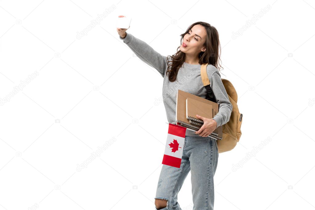 female student with notebooks holding canadian flag and taking selfie on smartphone isolated on white