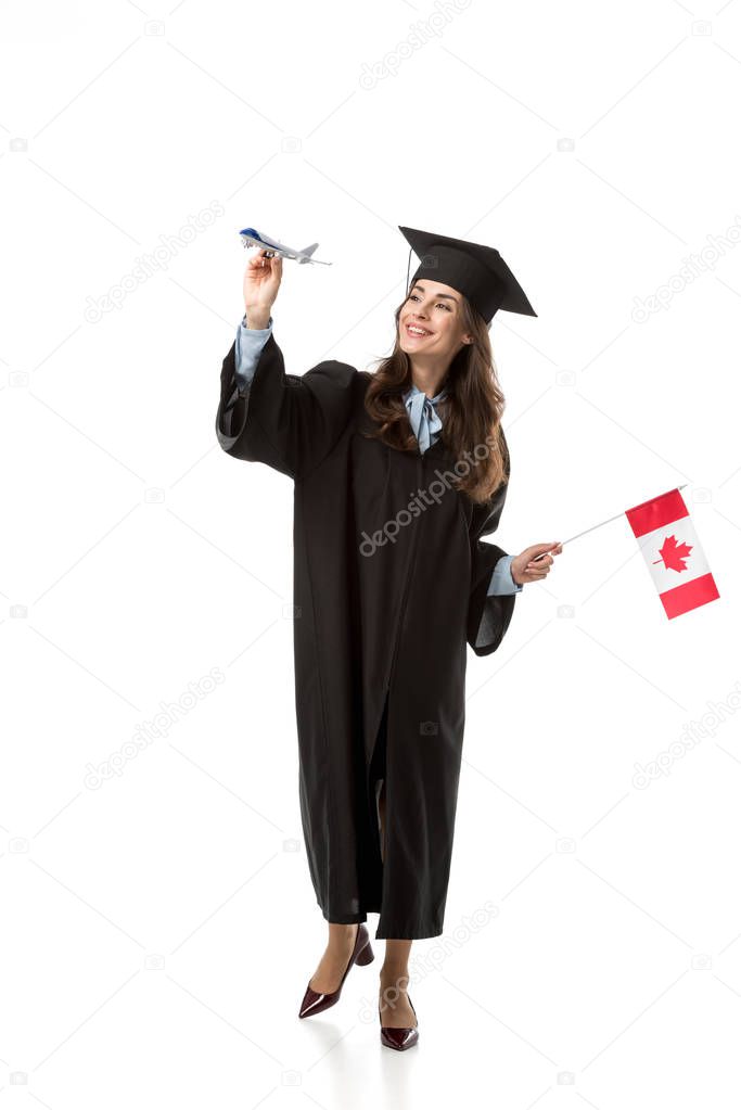 smiling female student in academic gown holding canadian flag and plane model isolated on white, studying abroad concept