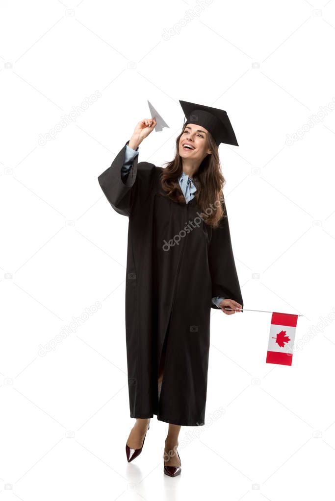 cheerful female student in academic gown holding canadian flag and paper plane isolated on white
