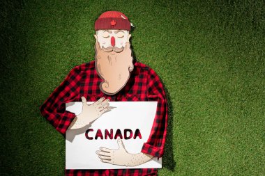 cardboard man in plaid shirt and hat holding white board with 'Canada' word on green grass background clipart