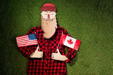 karton adam ekose gömlek ve Kanada ve Amerikan bayrakları yeşil çim zemin üzerine tutarak şapka