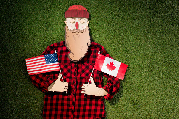 cardboard man in plaid shirt and hat holding canadian and american flags on green grass background