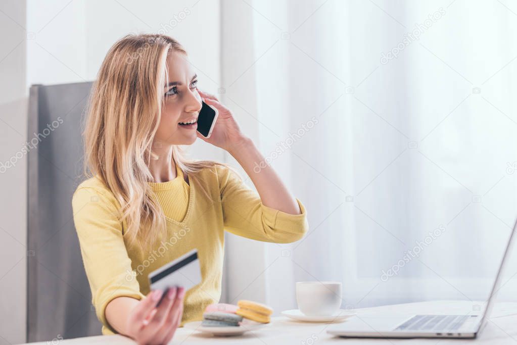 attractive blonde woman talking on smartphone while holding credit card near laptop