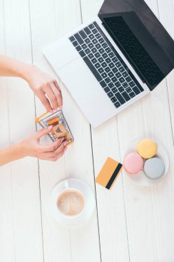 laptop, fincan kahve ve kurabiye yakınındaki küçük alışveriş arabası tutan üstten görünüm kadın