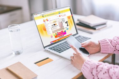 smartphone yakınındaki Aliexpress Web sitesi ile dizüstü bilgisayar ekranında tutan kadın görünümünü kırpılmış