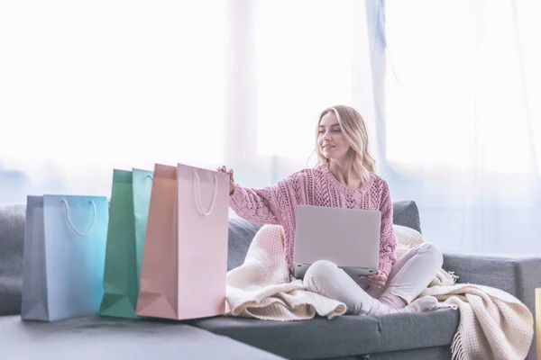 有吸引力的金发碧眼的女人坐在沙发上与笔记本电脑附近的购物袋 — 图库照片