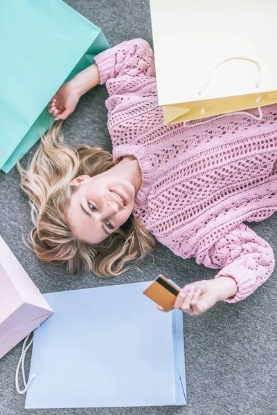 クレジット カードを見て 買い物袋の近くの床に横たわっている陽気な女性のトップ ビュー — ストック写真
