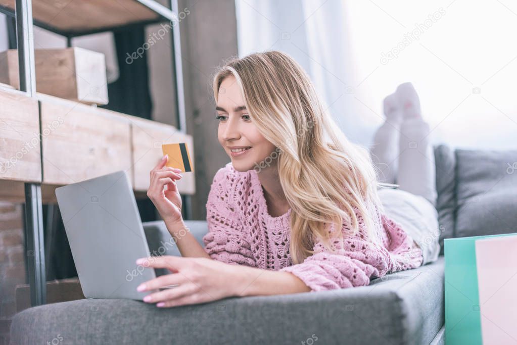 smiling woman holding credit card while lying on sofa with laptop