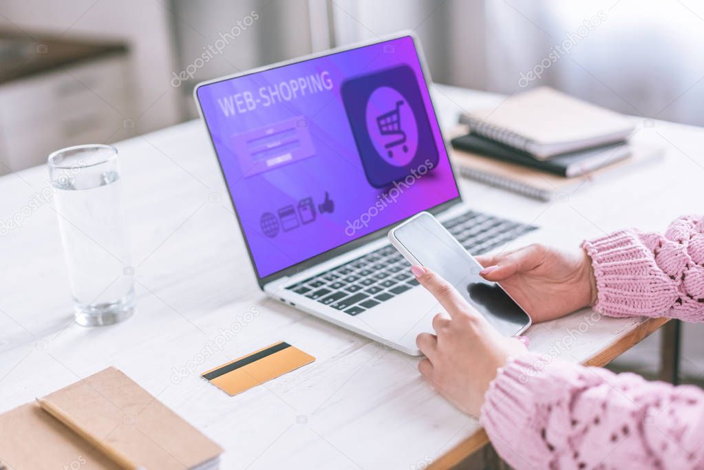 cropped view of woman holding smartphone near laptop with web shopping app on screen