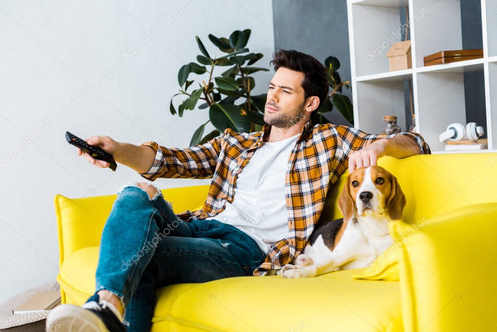 casual man with remote control watching tv and sitting on sofa with dog 