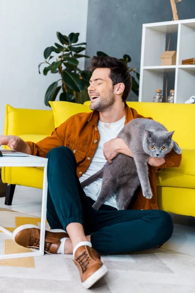 灰色の猫が付いている床に座ってラップトップを使用して若い笑い男 — ストック写真