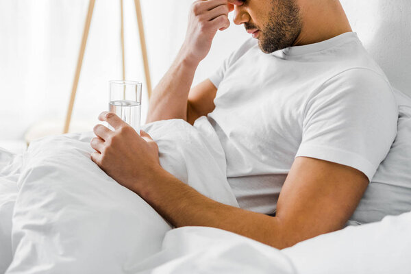 обрезанный вид человека с головной болью держа стакан воды в постели
 