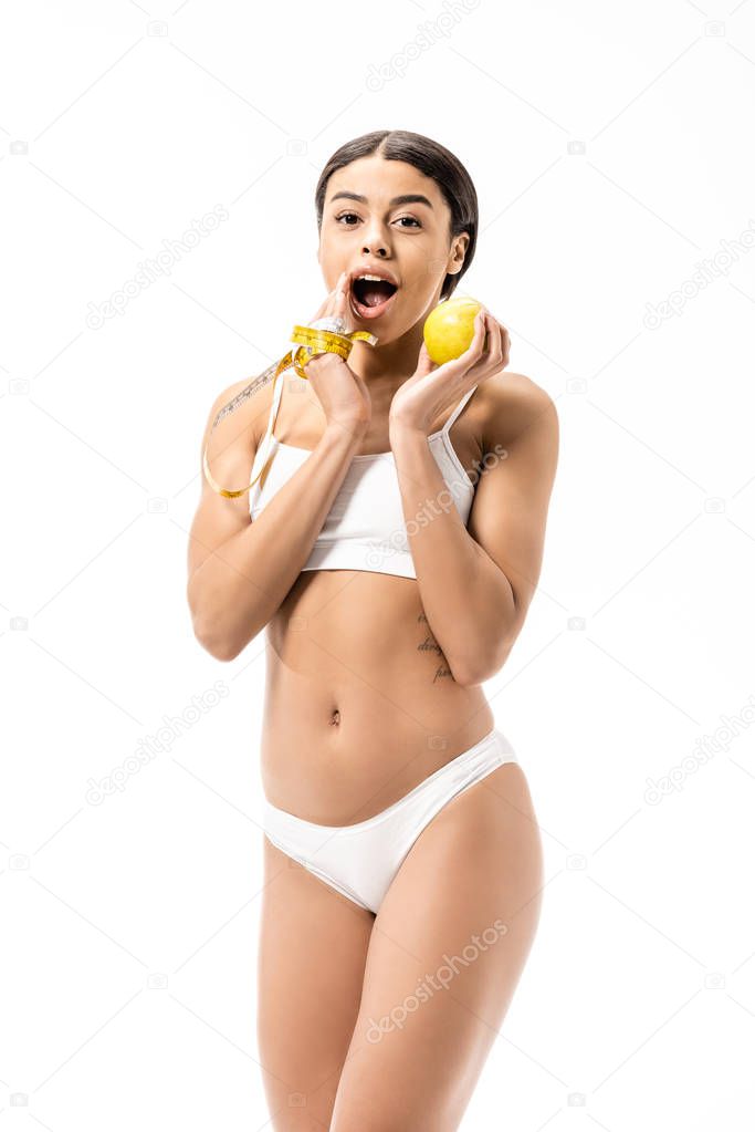 surprised young african american woman with open mouth holding apple and measuring tape isolated on white