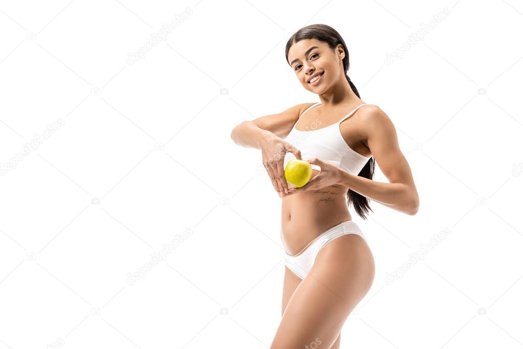 beautiful young african american woman holding green apple with hand heart symbol and smiling at camera isolated on white