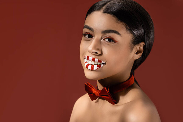 beautiful naked african american woman with red bow tie and creative makeup smiling at camera isolated on burgundy