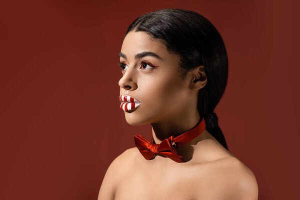 beautiful naked african american woman with red bow tie and creative makeup looking away isolated on burgundy