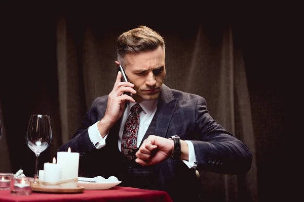handsome man in suit sitting at table and looking at watch while talking on smartphone in restaurant