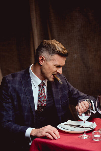 dissatisfied handsome man in suit sitting at table, smoking cigar and looking at watch in restaurant