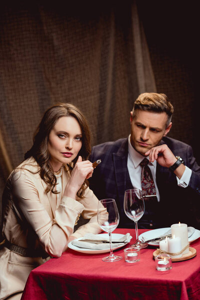 beautiful woman sitting at table and smoking cigar while having romantic dinner with handsome man in restaurant