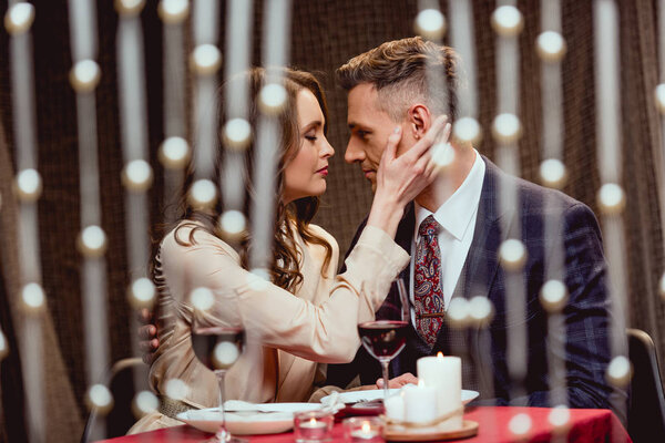woman gently touching face of man during romantic date in restaurant with bokeh lights on foreground
