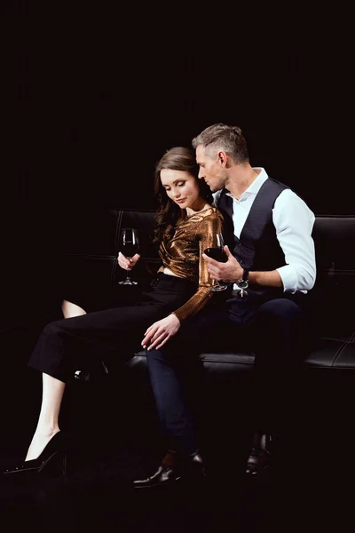 beautiful romantic couple sitting on couch with glasses of red wine isolated on black