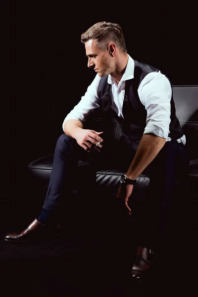 英俊的男人在正式穿坐在沙发上的香烟孤立的黑色 — 图库照片