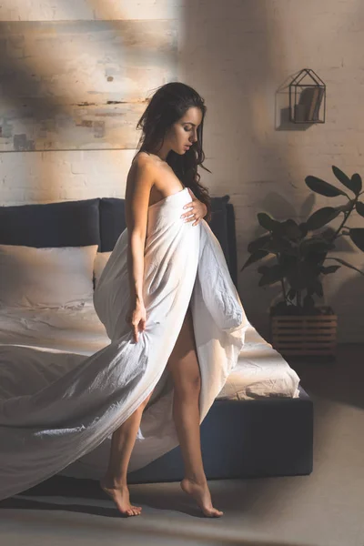 beautiful sensual woman covering body with duvet and standing on tiptoe in bedroom