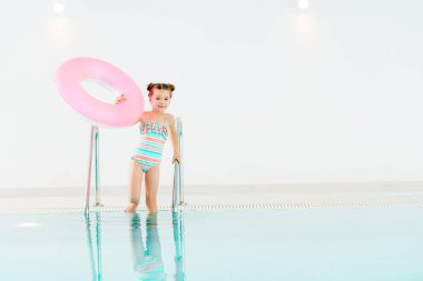 adorable kid standing in swimwear with inflatable ring and holding handrail in swimming pool