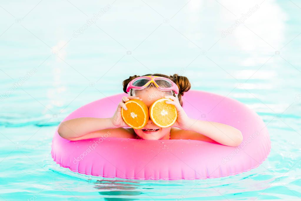 happy child holding half oranges near eyes while  swimming with inflatable ring in swimming pool