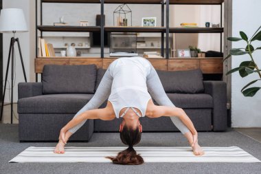 young woman practicing wide legged forward bend pose at home in living room clipart