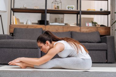 woman practicing seated forward fold pose on fitness mat at home in living room clipart