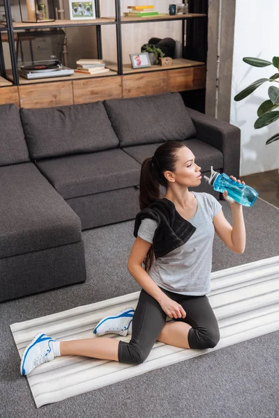 坐在健身垫上的女运动员 客厅里用运动瓶喝水 — 图库照片