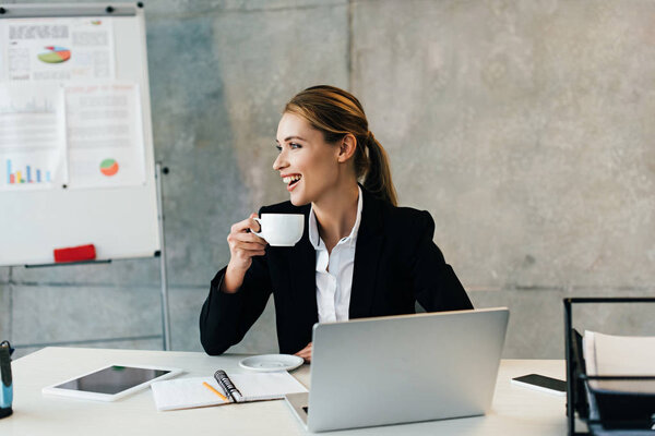 Beautiful laughing businesswoman sitting at workplace and drinking coffee 