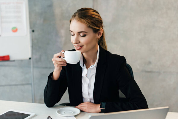 attractive businesswoman enjoys drinking coffee with closed eyes while sitting at workplace