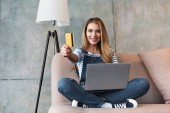 schöne Frau zeigt Kreditkarte und sitzt auf Sofa mit Laptop 