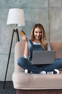 kredi kartı elinde tutan ve dizüstü bilgisayar ile kanepede oturan mutlu güzel kadın 