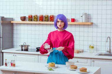 housewife with purple hair and colorful clothes looking at camera while preparing pancakes in kitchen