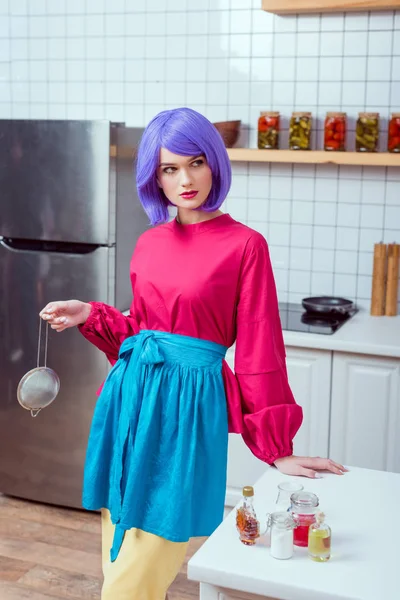紫髪とふるいを押しながらキッチンでポーズのカラフルな服を持つ美しい主婦 — ストック写真