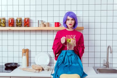 housewife with purple hair and colorful clothes sitting on kitchen counter and holding glass jar of lemonade