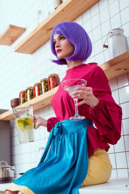 housewife with purple hair and colorful clothes sitting on kitchen counter and holding jar of lemonade with glass 