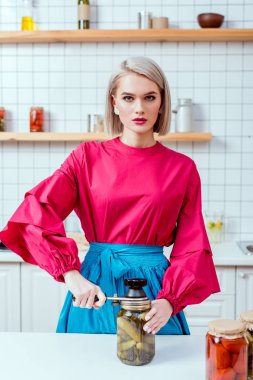 beautiful fashionable housewife sealing jar of pickled cucumbers in kitchen and looking at camera clipart