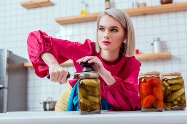 selective focus of beautiful fashionable housewife sealing jar of pickled cucumbers in kitchen  clipart