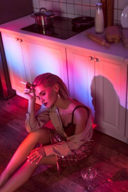 selective focus of beautiful fashionable woman sitting in kitchen and smoking in neon light