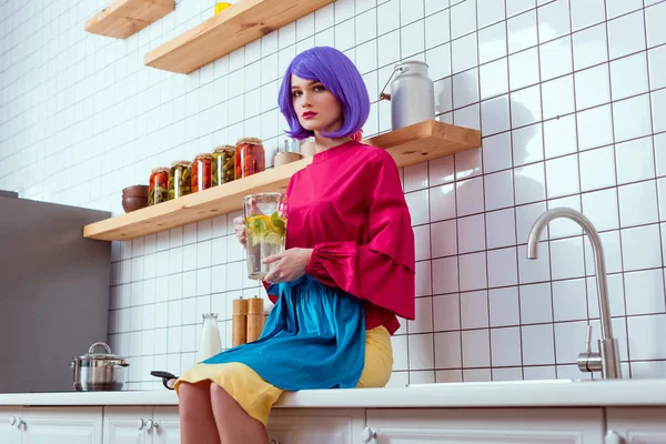 紫髪や服を着てカラフルなキッチン カウンターの上に座って レモネードのガラス瓶を保持している深刻な主婦 — ストック写真