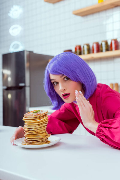 удивленная домохозяйка с фиолетовыми волосами и блинами, смотрящая в камеру и жестикулируя рукой на кухне
