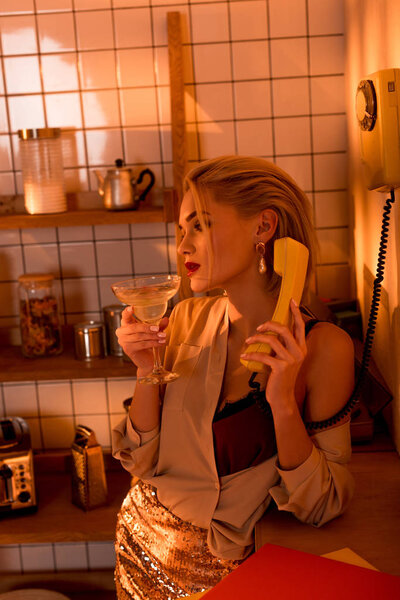 beautiful elegant woman with cocktail talking on retro telephone in kitchen with orange light