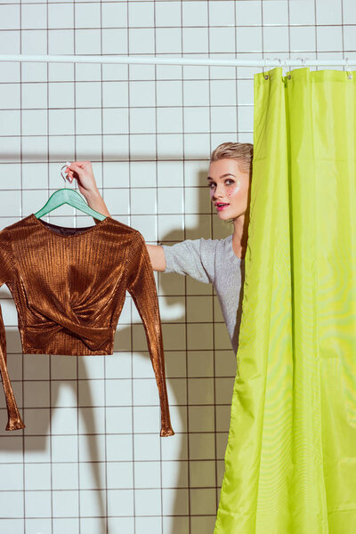 woman holding bronze top on hanger and looking at camera in shower with green curtain