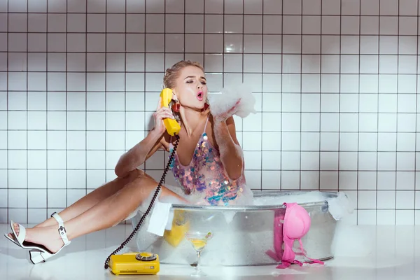 stock image beautiful young woman in wash tub talking on retro telephone and blowing foam bubbles in bathroom